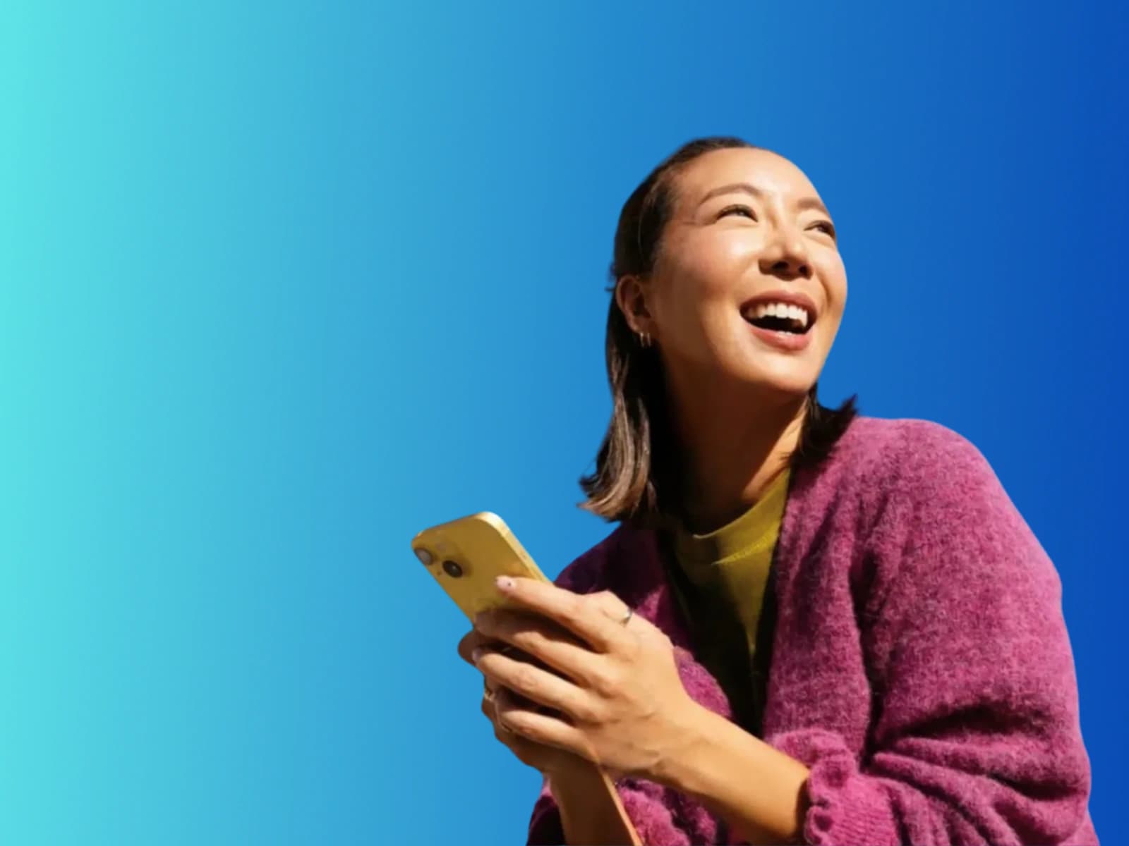 A smiling young woman holding her phone and looking up, against a bright blue sky